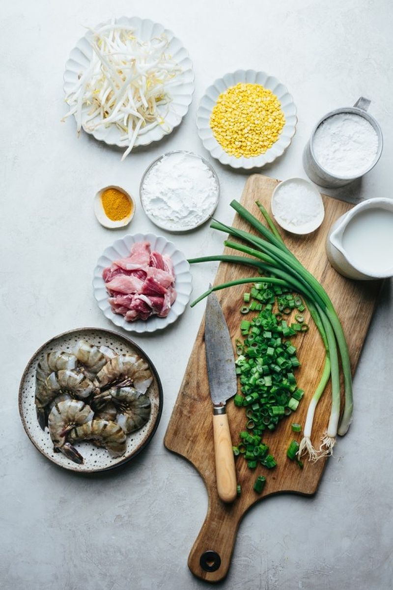 Ingredients for crepes: scallion on chopping board with knife, plate of prawns, plate of sliced pork, plate of turmeric, bowl of cornstarch, plate of mung bean, plate of bean sprouts, bowl of salt, cup of rice flour, and cup of coconut cream